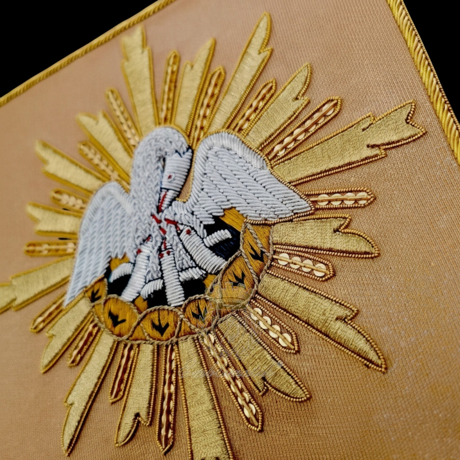 Emblematik by Pasamanerias y Bordados hand-embroidered pelican with metal wire threads on tan fabric, surrounded by gold sunburst.